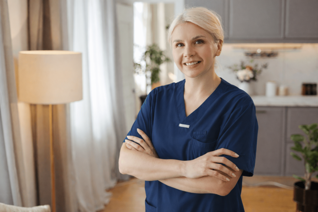 Pflegefachkraft in blauer Berufskleidung steht lächelnd mit verschränkten Armen in einer wohnlichen Umgebung – professionelle Pflegekraft im häuslichen Pflege- oder Betreuungsumfeld.